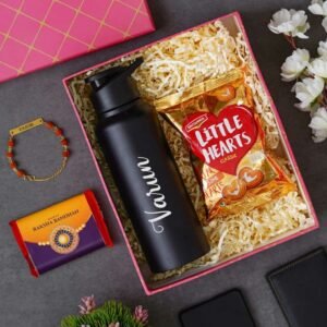 Personalized Rakhi gift box containing a custom black water bottle with the name Varun, a packet of Little Hearts biscuits, a Rudraksha Rakhi, and a Raksha Bandhan-themed chocolate bar, displayed in a pink and gold gift box with decorative items.