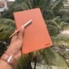 Customized orange leatherette journal held in hand with personalized pen, engraved with the name Rahul, against a backdrop of palm trees.
