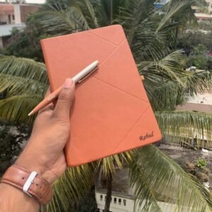 Customized orange leatherette journal held in hand with personalized pen, engraved with the name Rahul, against a backdrop of palm trees.
