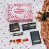 Valentine's Day themed men's gift box with customized accessories including a wallet, passport cover, sunglass case, keychain, and rose chocolate, adorned with your name