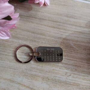 Real photo of personalised drive safe metal calendar keychain front side with custom May 2025 date engraving and 18 circled with heart, styled on wooden surface with pink flowers