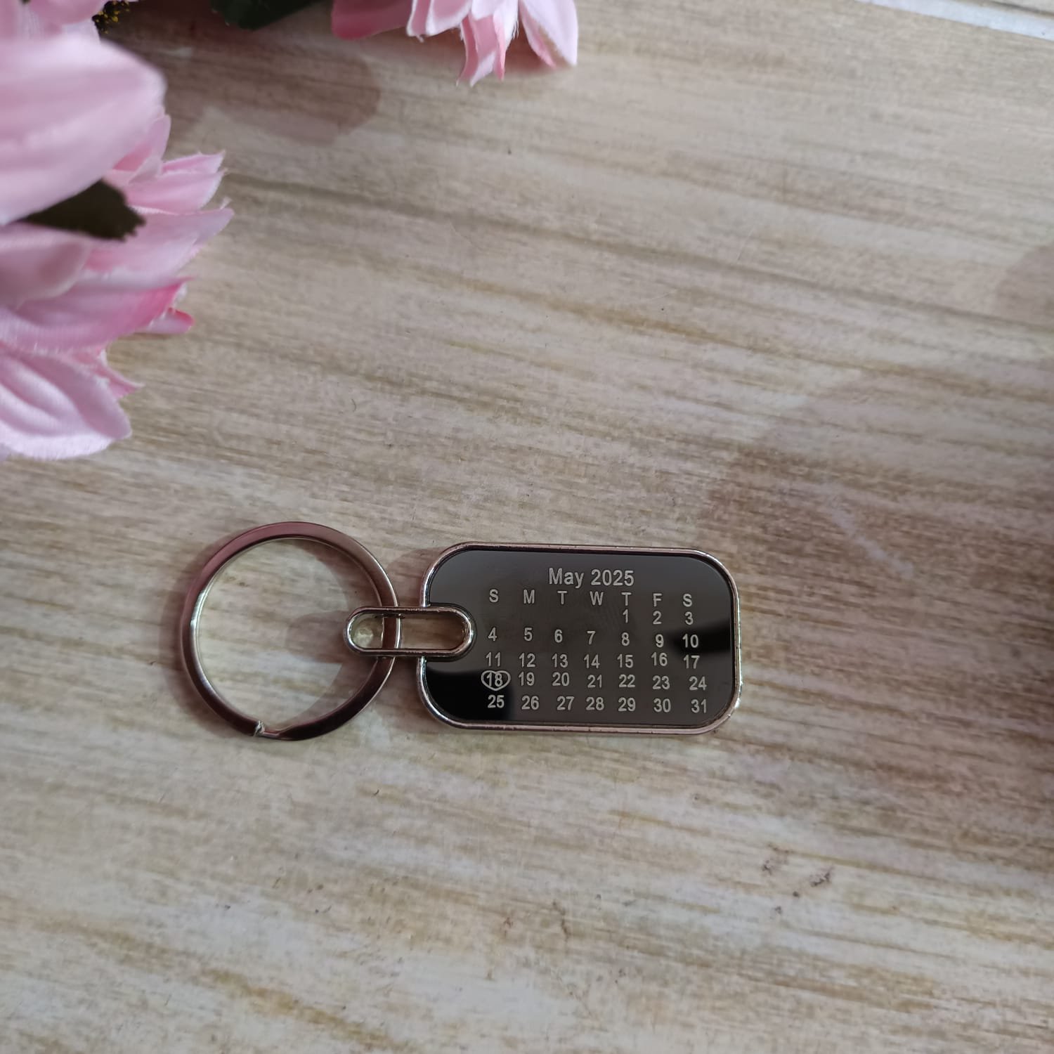 Real photo of personalised drive safe metal calendar keychain front side with custom May 2025 date engraving and 18 circled with heart, styled on wooden surface with pink flowers